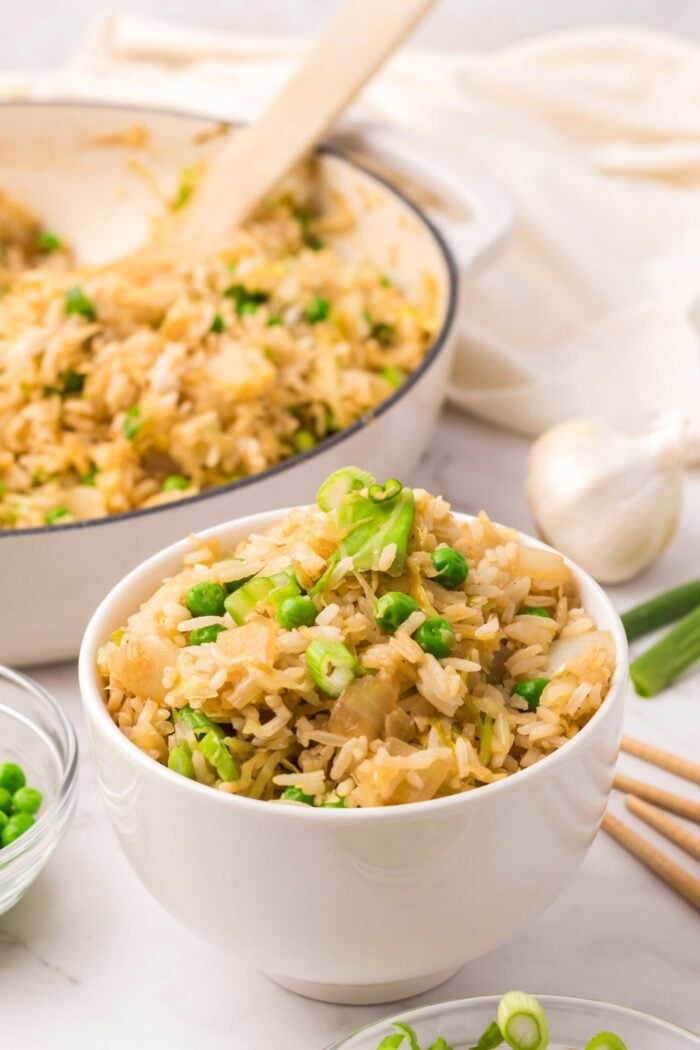 A bowl of cabbage fried rice with green peas and green onion beside a large skillet of fried rice with a wooden spatula in it.
