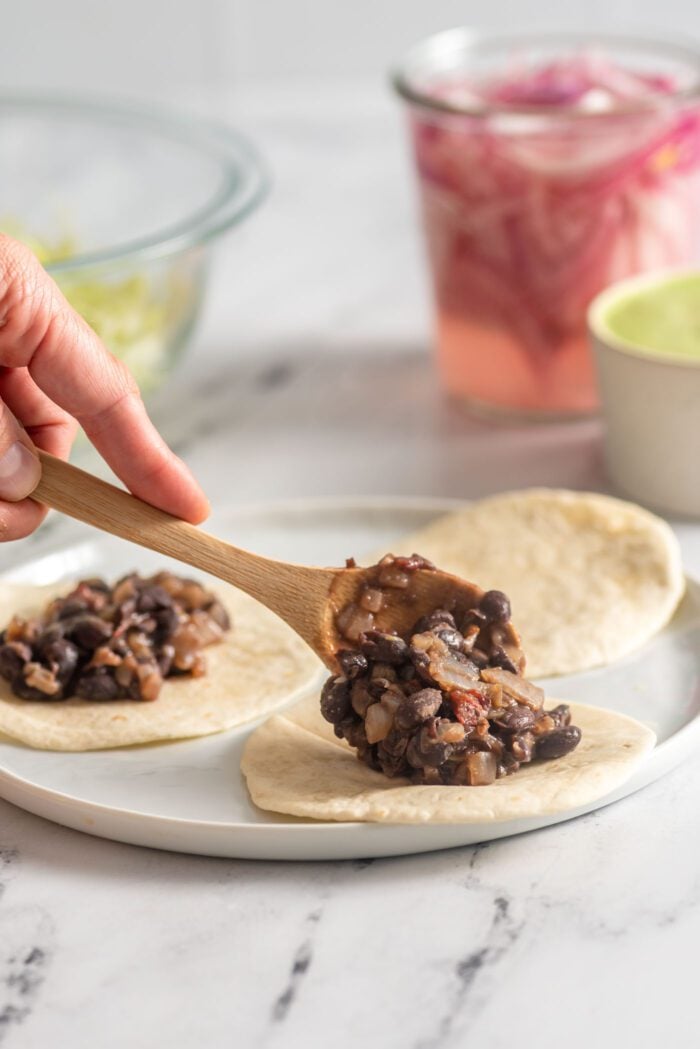 Adding a scoop of black beans to a small soft taco shell..