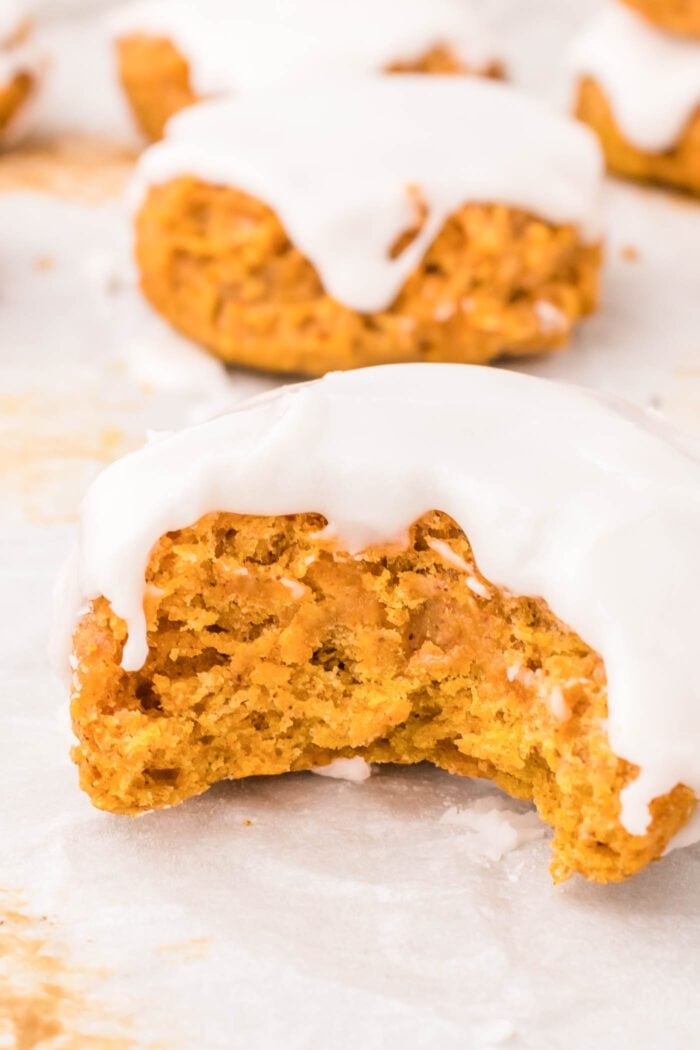 Close up of a soft pumpkin cookie with glaze with a bite taken out of it so you can see the texture inside. The cookie is sitting on a baking tray lined with parchment paper and there are more cookies in the background.