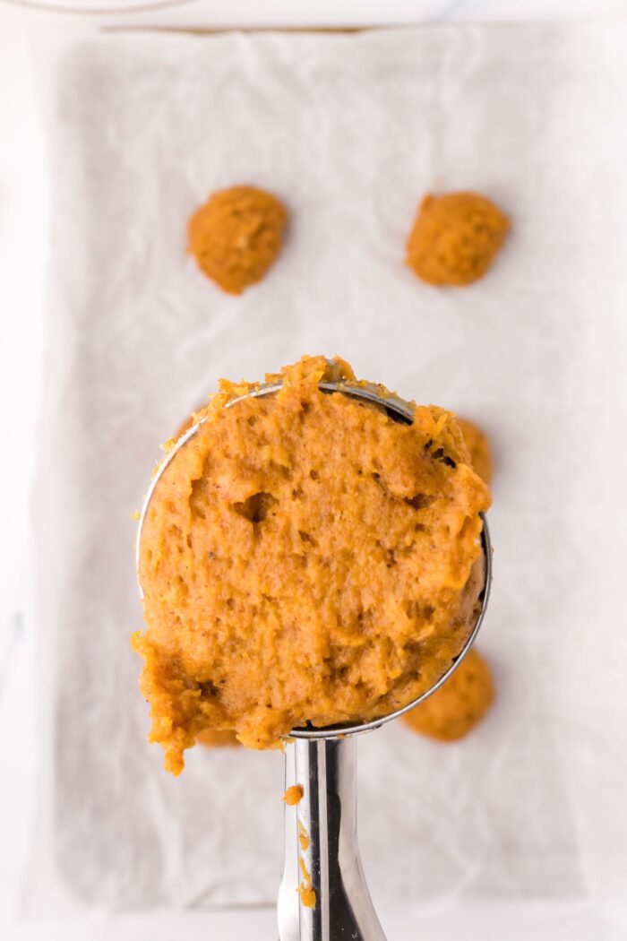 Pumpkin cookie dough in a cookie scoop held over a baking tray of raw cookies.