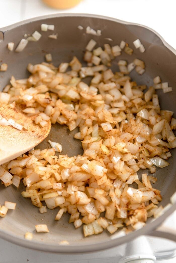 Diced onions with spices cooking in a skillet with a wooden spoon.