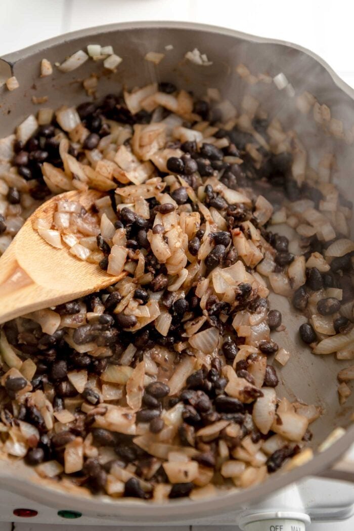 Black beans and onions cooking with spices in a skillet. The mixture is steaming and there's a wooden spoon resting in the skillet.