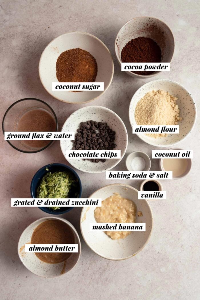 All the ingredients needed for a zucchini brownie recipe gathered in various bowls. Each ingredient is labelled with text overlay.
