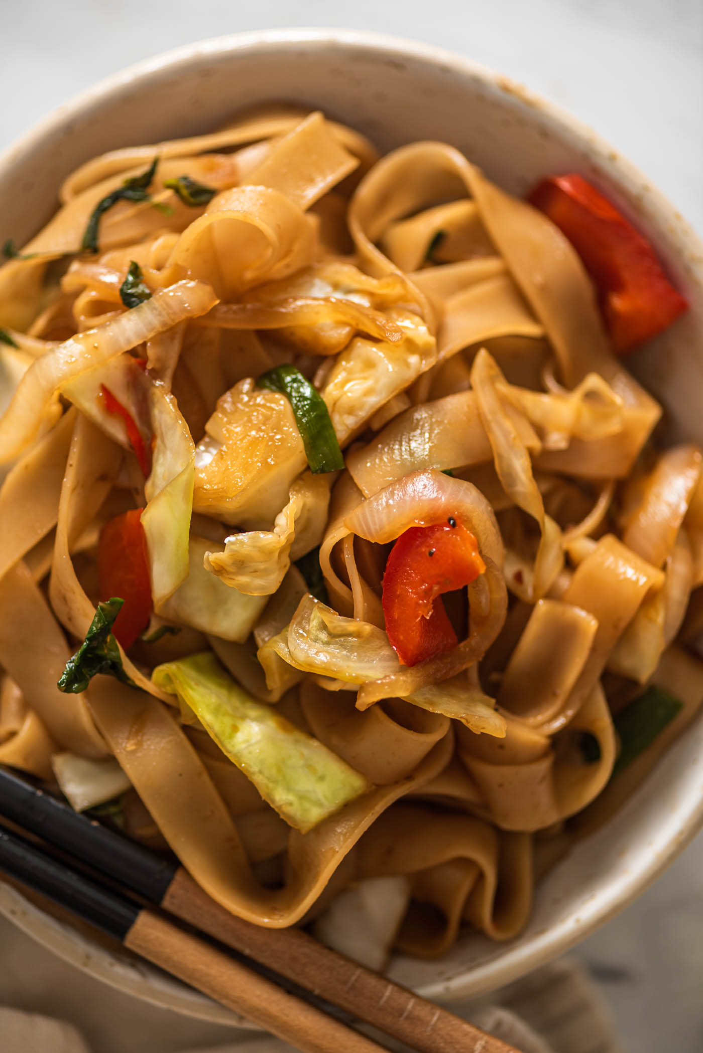 A bowl of drunken noodles with veggies. A set of chopsticks rests on the side of the bowl.
