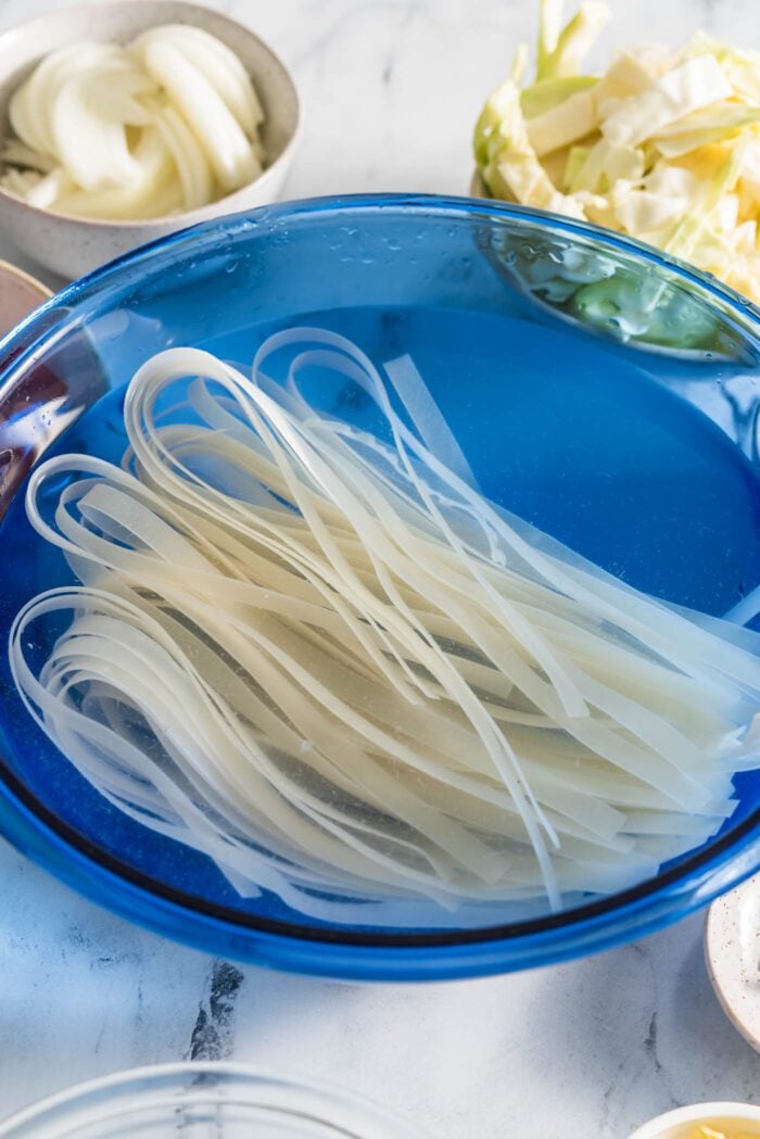 Wide rice noodles soaking in a large glass bowl.