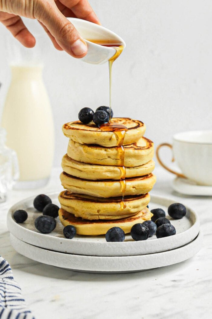 Hand drizzling maple syrup from a small dish over a stack of chickpea pancakes on a plate. The pancakes are also topped with blueberries.