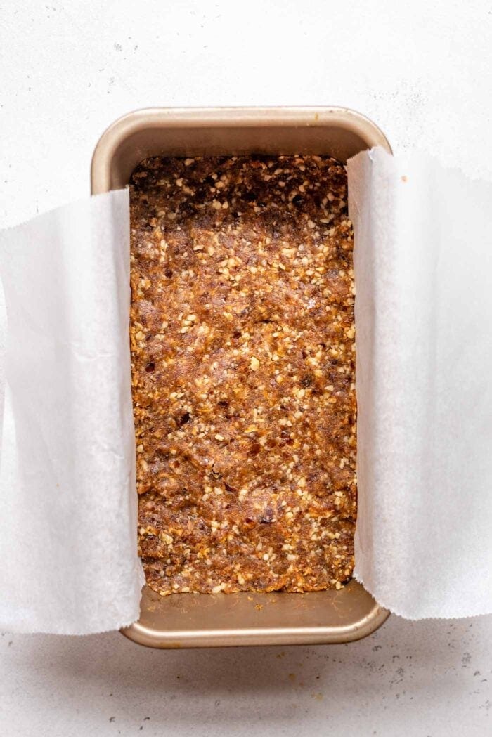 A raw walnut date crust pressed into a loaf pan lined with parchment paper.
